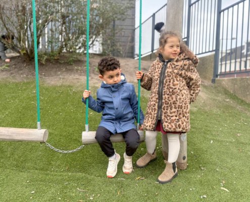 Twee kinderen spelen samen bij de schommel waarvan er een kind op de schommel zit en de ander er naast staat.