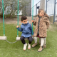 Twee kinderen spelen samen bij de schommel waarvan er een kind op de schommel zit en de ander er naast staat.