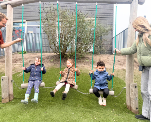 Drie kinderen zitten vrolijk te schommelen op de schommel met aan beide zijden van de schommel een begeleider.