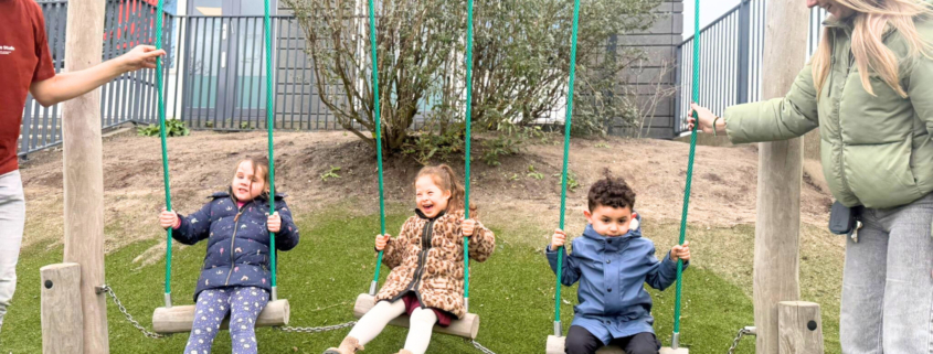Drie kinderen zitten vrolijk te schommelen op de schommel met aan beide zijden van de schommel een begeleider.