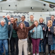 Groepsfoto van diverse leden van de Jostiband op een rij voor een millitaire helikopter. Zij juichen en steken hun handen in de lucht van blijdschap.